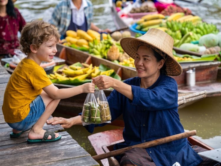 Young boy buying a bag of Thai sweets from a smiling vendor in a straw hat on her fruit boat at Damnoen Saduak Floating Market near Bangkok